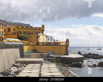 Blick auf das gelbe Fort Sao Tiago aus dem 17. Jahrhundert in funchal madeira, vom Meer mit sonnendurchflutetem blauem Meer und Booten mit der Hügelsurro Stockfoto