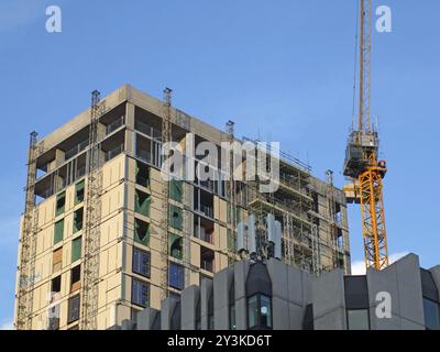 Ein hoher gelber Baukran, der an einem großen, hochgelegenen Betongebäude mit Gerüsten vor blauem Himmel arbeitet Stockfoto