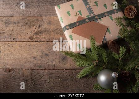 Weihnachtsgrußkartenrahmen mit einem braunen Geschenk mit grünen Bäumen, umgeben von Tannenzweigen, Tannenzapfen und Weihnachtskugeln auf einem alten Tisch Stockfoto