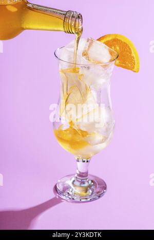 Zubereitung eines kalten Sommergetränks, indem Orangensirup über Eiswürfel in einem Glas auf violettem Hintergrund gegossen wird. Orangenlimonade machen. Tropische Mocktail Stockfoto