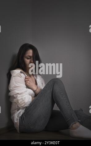 Die junge Frau drückte sich deprimiert, zog ihr Hemd, um sich zu bedecken, während sie auf dem Boden saß und sich an eine Wand lehnte Stockfoto