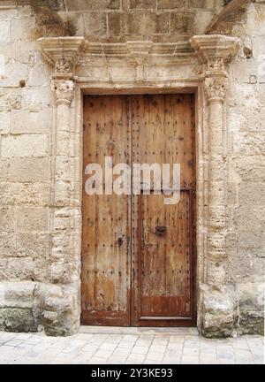 Eine große alte braune Holztür, die mit rostigen Eisenbolzen bedeckt ist, Schlüsselloch und Griff in einem kunstvoll geschnitzten Steinrahmen mit umliegender Wand Stockfoto