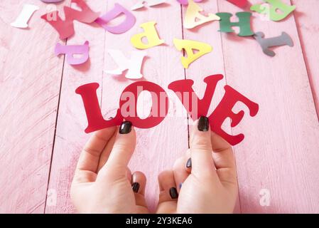 Frauenhände mit schwarzen Nägeln, die das Wort Liebe aus roten Papierbuchstaben über einem rosafarbenen Holztisch halten, mit bunten Buchstaben im Hintergrund Stockfoto