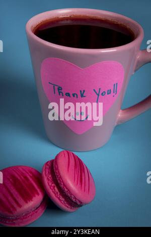 Vielen Dank Nachricht handschriftlich auf einem Herzförmigen Haftnotiz. Rosa Becher Kaffee und zwei Rosa Makronen auf einer blauen Tabelle Stockfoto