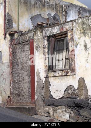 Ein altes Ruinenhaus brach teilweise an einer schrägen Straße zusammen, mit einer versperrten Tür, zerbröckelten Wänden und verblassten rot lackierten Fenstern mit geschlossenen Fensterläden Stockfoto