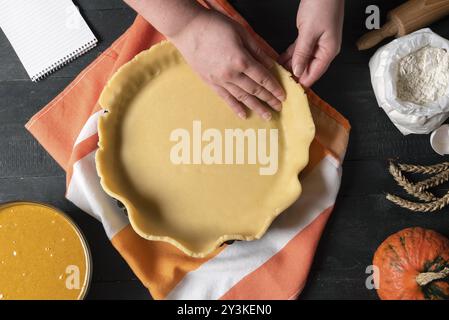 Frau legt die Kuchenkruste auf ein Backblech, auf ein Küchentuch, und Kürbiskuchen-Füllung, Mehl, Nudelnadel, auf einen rustikalen Tisch. Ferienhaus backen Stockfoto