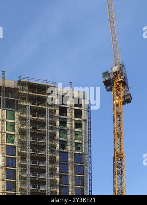 Ein hoher gelber Baukran, der an einem großen, hochgelegenen Betongebäude mit Gerüsten vor blauem Himmel arbeitet Stockfoto