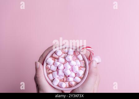 Weihnachtsgetränk, eine Tasse heiße Schokolade mit rosa Mini-Marshmallows und einem Weihnacht-Zuckerrohr, gehalten mit zwei Händen, auf einem rosa Hintergrund Stockfoto