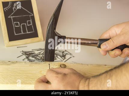 Bild mit der Hand eines Baumeisters, der einen kleinen Nagel in ein Holzbrett schlägt, und im Hintergrund eine Tafel mit einer lustigen Zeichnung eines Hauses Stockfoto