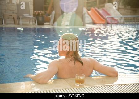 Ein Mann, der sich zurück am Rand des Pools lehnt, hinter ihm Drink platziert, einen friedlichen, sonnendurchfluteten Moment im Pool genießt Stockfoto