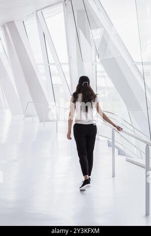 Eine junge Frau, ungezwungenermaßen gekleidet, geht auf einem hellen, weißen Flur von einem Bürogebäude mit hohen Fenstern und modernen Säulen aus Stockfoto