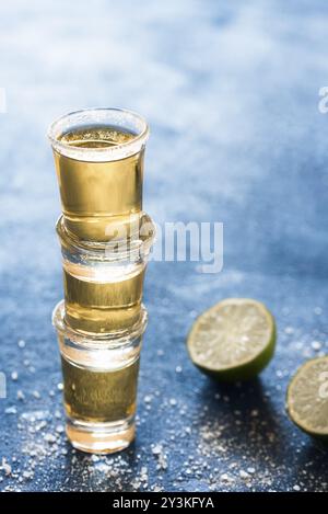 Drei Schüsse Tequila gestapelt auf einem blauen Tisch. Goldenes Tequila-Glas mit Limette und Salz. Aromatischer starker mexikanischer Alkohol. Partyaufnahmen Stockfoto