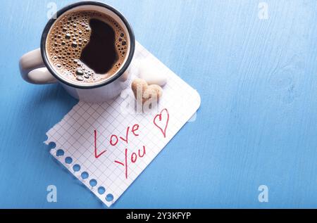 Niedliches romantisches Bild mit einer Tasse heißem Kaffee, herzförmigem Zucker und einer Papiernotiz mit Love You Botschaft auf einem blauen Holztisch Stockfoto