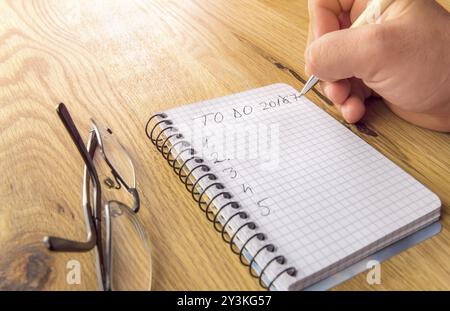 Die Hand des Mannes schreibt die To-do-Liste für 2017, auf einem Spiralnotizbuch, auf einem Holztisch mit Brille Stockfoto