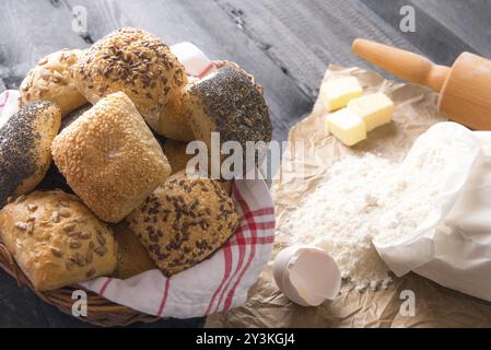 Backmotiv mit einem Haufen gemischter Brötchen, in einem Korb, mit Mehl, Eiern und Butter in der Nähe, auf schwarzem Holzhintergrund Stockfoto