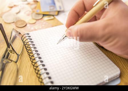 Nahaufnahme mit einem leeren Mathematiknotizbuch und einer Hand, die einen Stift hält. Im Hintergrund stehen Geld, Karten, Scheine und eine Brille Stockfoto