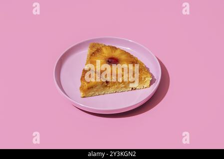 Eine Scheibe Ananaskuchen auf einem rosafarbenen Teller minimalistisch auf einem farbenfrohen Tisch. Hausgemachter umgedrehter Kuchen mit einer Ananasscheibe Stockfoto