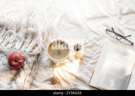 Gemütliches Freizeitkonzept mit offenem Buch, Gläsern, einem Apfel auf einem flauschigen Tuch und einer heißen Tasse Kaffee, auf einer weißen Decke, in der Morgensonne Stockfoto