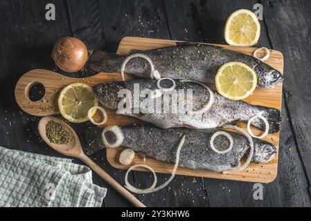 Blick aus einem hohen Winkel mit ungekochtem Fisch auf einem hölzernen Schneidebrett, abgeschmeckt mit getrockneten Kräutern, Zitronenscheiben und Zwiebelringen, auf einem Vintage schwarzen Tisch Stockfoto