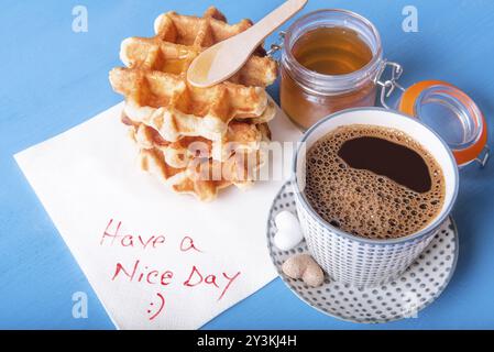 Eine Tasse Kaffee mit herzförmigem Zucker, hausgemachte Waffeln mit Honig und eine Serviette mit einer schönen Tagesbotschaft auf einem blauen Holztisch Stockfoto