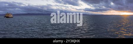 Panorama mit Sonnenuntergang über dem Seewasser und Einzelboot Segeln mit Passagieren, auf dem Bodensee in der Stadt Friedrichshafen, Deutschland, Europa Stockfoto