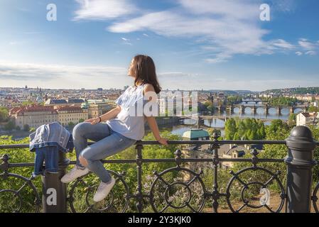 Panorama der Prager Stadt, der Moldau mit ihren vielen Brücken und einer jungen, schönen Frau, die auf einem Zaun sitzt und die Sonne und die Aussicht genießt Stockfoto