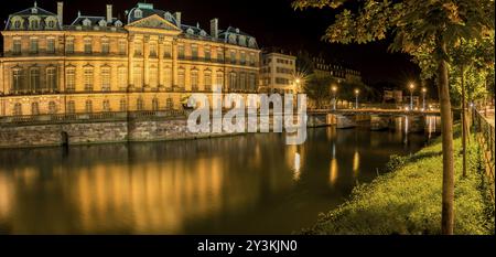 Nächtlicher Panoramablick auf den Rohan Palast und den Fluss Ill, mit Gebäuden und gelben Lichtern, die sich im Wasser spiegeln Stockfoto