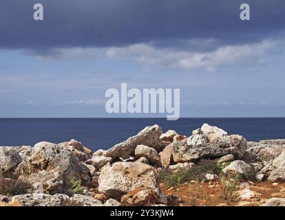 Eine felsige Küstenszene mit weißen Steinen vor einem ruhigen blauen Meer und Himmel mit Sonnenlicht und Wolken Stockfoto