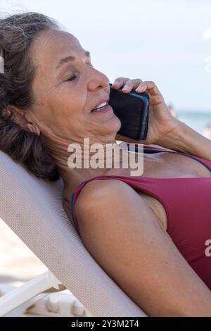 Eine ältere Frau liegt auf einer Liege am Strand und führt ein Gespräch mit ihrem Smartphone. Stockfoto