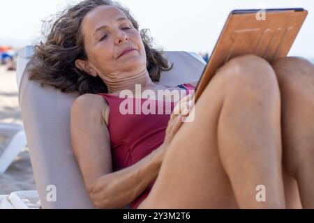 Eine ältere Frau liegt bequem auf einem Liegestuhl, liest oder schaut sich ein Tablet an. Stockfoto