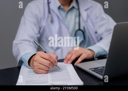 Doktor schreibt Dokumente, mit Laptop. Ausfüllen medizinischer Unterlagen, auf Genauigkeit ausgerichtet. Professionelle Gesundheitspraxis, Klinikumgebung. Medi Stockfoto