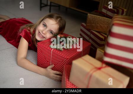 Das aufgeregte Kind in rotem Kleid umarmt fröhlich das Weihnachtsgeschenk zu Hause Stockfoto