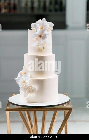 Eleganter dreistöckiger Hochzeitstorte mit zarten weißen Orchideen und goldenem Monogramm auf einem modernen geometrischen Standfuß. Stockfoto