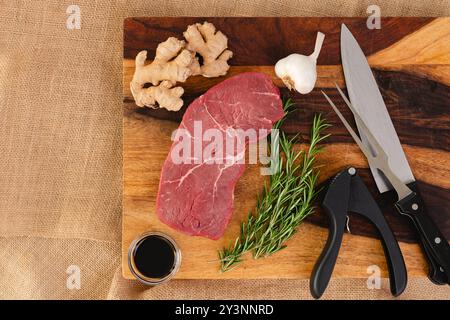 Rohe Rindersteak mit Ingwerwurzel, Knoblauch, Sojasauce und Rosmarin in Nahaufnahme auf einem hölzernen Schneidebrett, flach gelegt Stockfoto