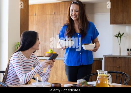 Zu Hause, serviert Essen, asiatische Mutter lächelt Tochter mit Smartphone in der Küche Essbereich an Stockfoto