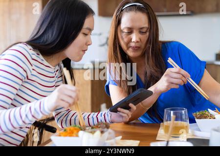 Zu Hause, mit Essstäbchen, asiatische Mutter und Tochter schauen beim gemeinsamen Essen auf das Smartphone Stockfoto