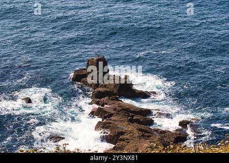 Ein Felsvorsprung im Ozean mit Wellen, die gegen ihn krachen. Das Wasser ist tiefblau, und die Szene fängt die natürliche Schönheit der Küstenlandschaft ein. Stockfoto