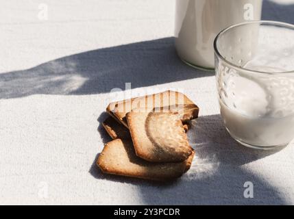 Ein Set flacher, hausgemachter Kekse, die neben einem Glas Milch auf einer weißen Tischdecke platziert werden, mit sanftem Sonnenlicht, das Schatten wirft, was ein ruhiges und rustikales Ou schafft Stockfoto