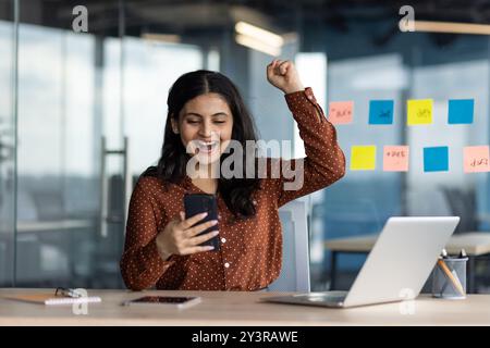 Joyful successful business woman at workplace holding phone in hands received notification of victory and hearing results of financial activity, Hispanic woman holding hands up triumph gesture. Stockfoto