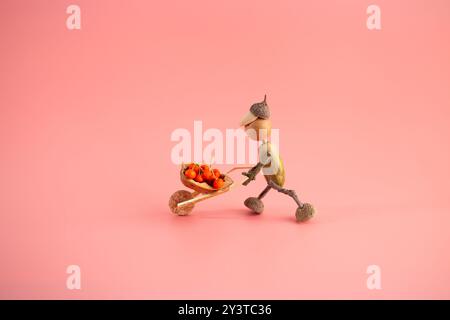 Eine Figur aus Eicheln, die eine Schubkarre voller Beeren drückt, handgefertigte Konzeptkunst, DIY-Herbstbastelidee für Kinder, lustige Aktivitäten, kreative Natur Stockfoto