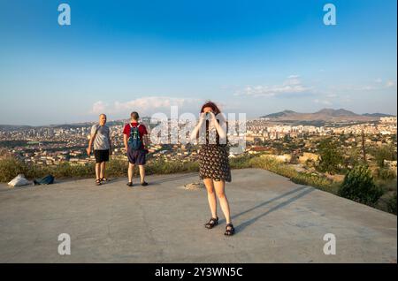 Eine Frau fotografiert mit ihrer Kamera ein malerisches Stadtbild, während zwei Männer den Panoramablick hinter ihr genießen, ein Fotograf in einem Blumenkleid umrahmt sie Stockfoto