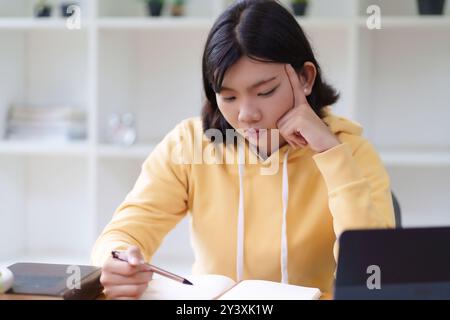 Eine junge Frau mit gelbem Hoodie sitzt an einem Tisch und schreibt in ein Notizbuch. Sie scheint konzentriert und tief in Gedanken zu sein, umgeben von einem sauberen und Stockfoto