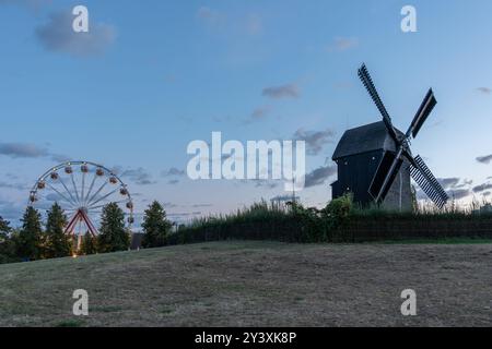 Eine Windmühle befindet sich im Hintergrund eines Feldes mit einem Karussell im Vordergrund. Der Himmel ist bewölkt und die Sonne untergeht Stockfoto