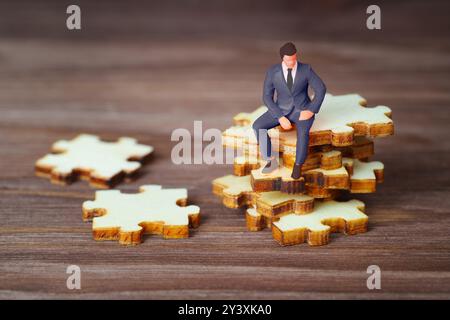 Miniaturfigur eines Geschäftsmannes, der auf Puzzleteilen sitzt und Problemlösung und strategisches Denken im Geschäft veranschaulicht. Stockfoto