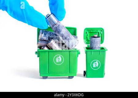 Hand mit Handschuhen, die stark korrodierte Batterien in grüne Miniatur-Recyclingbehälter legen, isoliert auf weißem Hintergrund. Sichere Entsorgung und umweltfreundliche bzw. Stockfoto