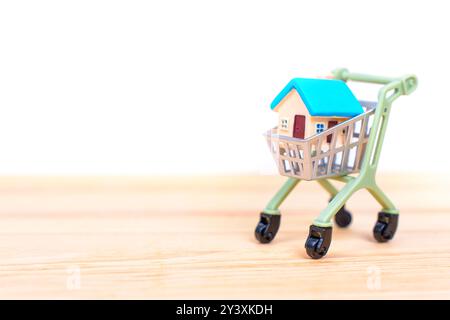 Hausmodell in einem Einkaufswagen auf hölzernem Hintergrund, das Konzepte des Wohnungskaufs und der Immobilieninvestition veranschaulicht. Stockfoto