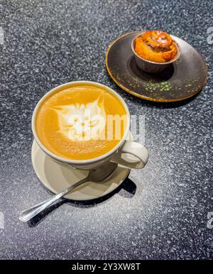 Kunstvoller Kaffee-Latte mit Katzengesicht auf der Schaumplatte, serviert auf einem Tisch mit frisch gebackenem Gebäck auf einem dunklen Teller. Hochwertige Fotos Stockfoto