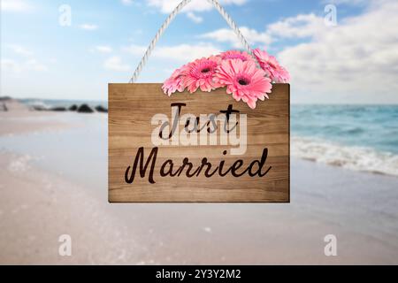 Flitterwochen. Holzbrett mit Worten, die gerade geheiratet wurden, und Blumen am Sandstrand Stockfoto