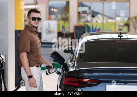 Gutaussehender Mann in Sonnenbrille, der sein Auto an der Selbstbedienungstankstelle betankt. Männlich betankt Diesel mit Benzin und sieht in die Kamera Stockfoto