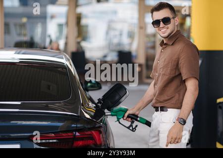 Lächelnder Mann, der sein Luxusauto an der Tankstelle betankt. Der Kerl guckt in die Kamera und gießt Benzin in den Tank eines modernen Fahrzeugs an der Tankstelle in der Stadt Stockfoto